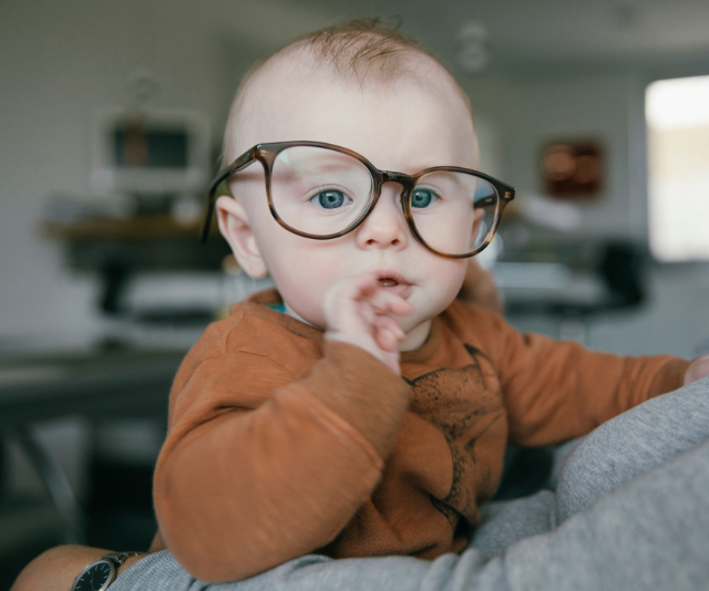 Adorable fair haired baby wearing brown onesie and wearing oversized adults reading glasses.