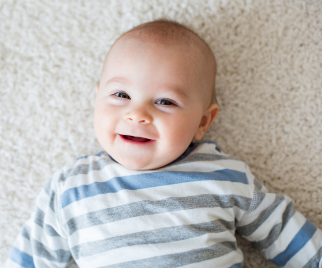 Cute baby boy smiling wile wearing a blue and white striped shirt while laying on the floor.