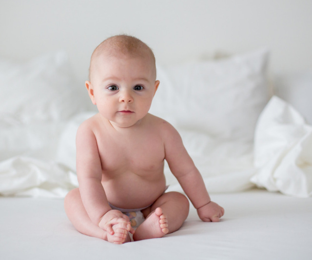 Adorable bald baby boy sitting up on a bed with white linen while wearing only a nappy.